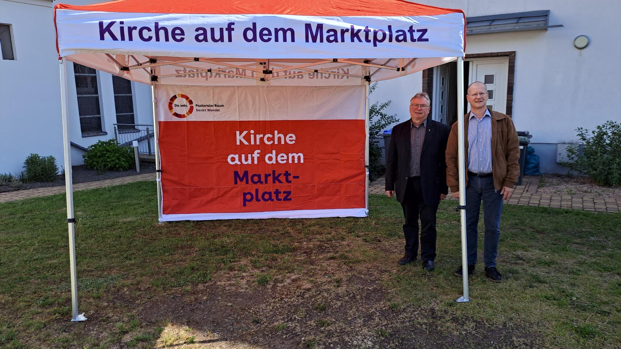 Man sieht ein Faltzelt und zwei Menschen, im Hintergrund ein Gebäude.