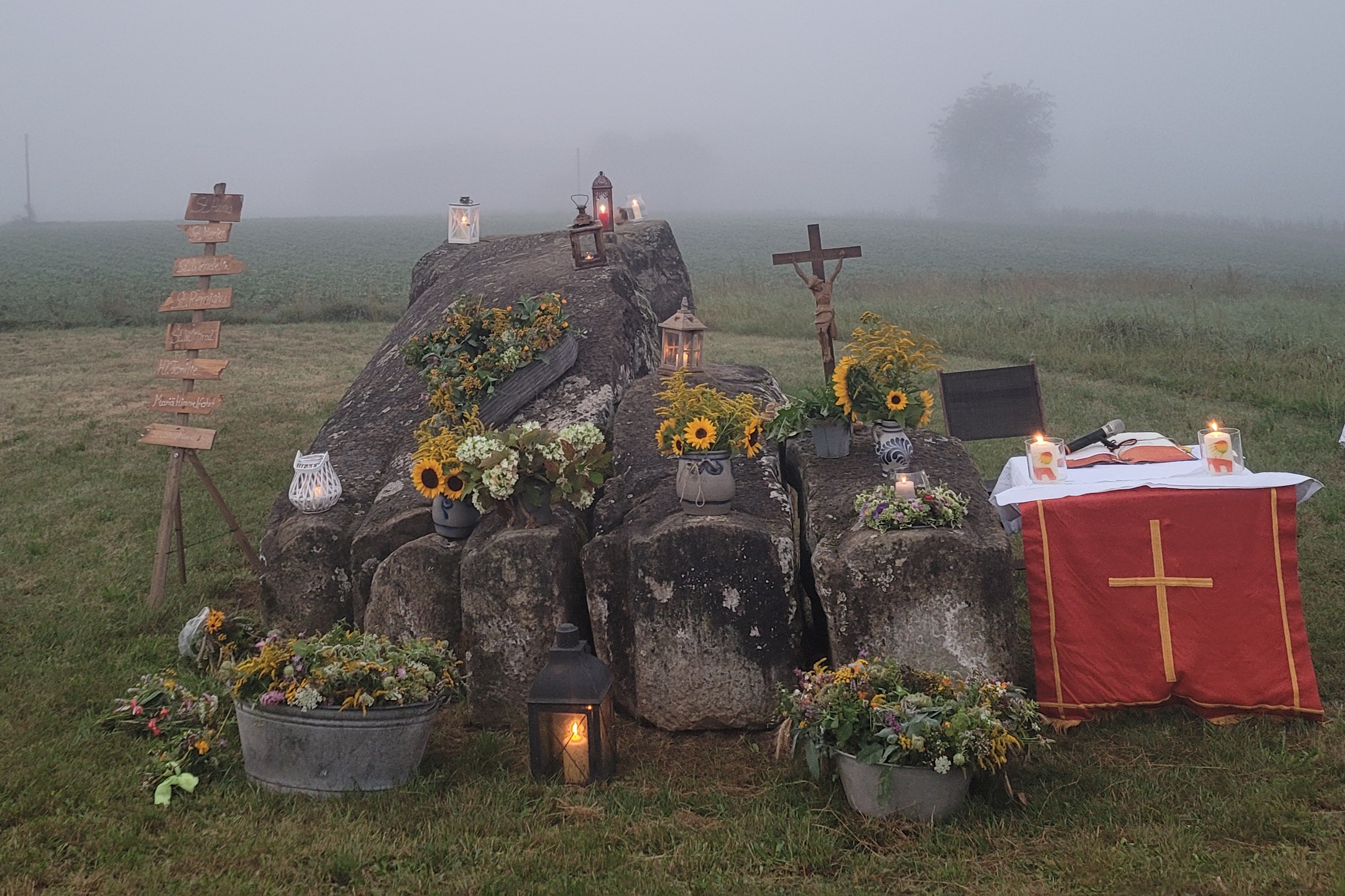 Sie sehen einen mit Blumen und Kerzen geschmückten Felsen neben einem kleinen Altar und ein Wegweiser. (c) Andrea Johann-Schneider Sie sehen einen mit Blumen und Kerzen geschmückten Felsen neben einem kleinen Altar und ein Wegweiser.