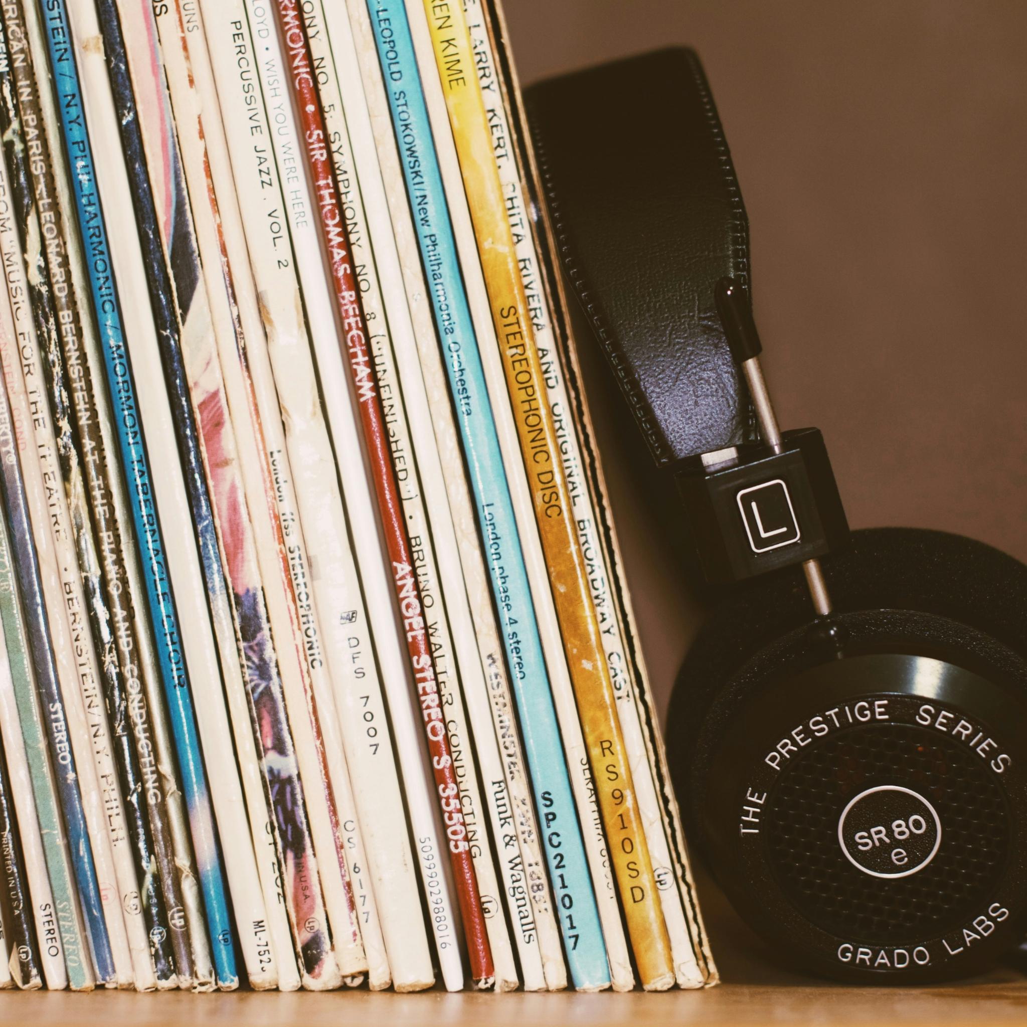 Kopfhörer, Schalplatten in einem Regal (c) https://unsplash.com/de/fotos/kabellose-kopfhorer-die-sich-an-bucher-anlehnen-T3mKJXfdims Kopfhörer, Schalplatten in einem Regal