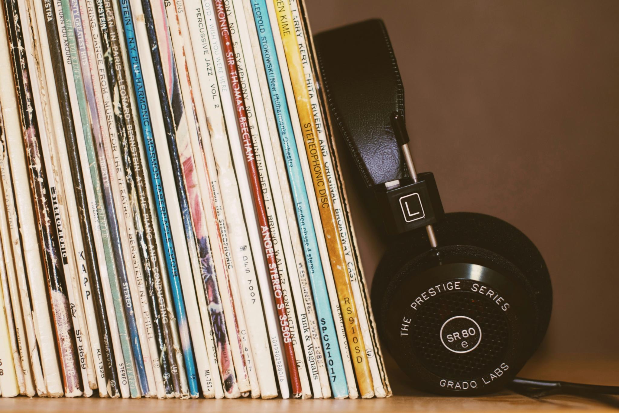 Kopfhörer, Schalplatten in einem Regal (c) https://unsplash.com/de/fotos/kabellose-kopfhorer-die-sich-an-bucher-anlehnen-T3mKJXfdims Kopfhörer, Schalplatten in einem Regal