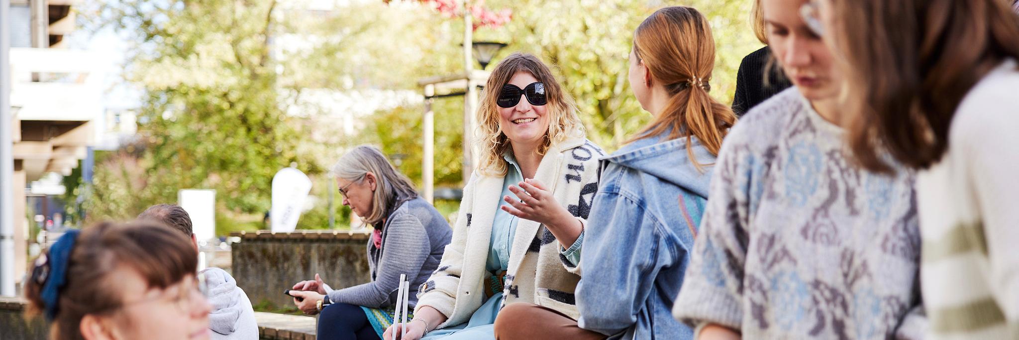 Mehrere Frauen sitzen auf stufenförmig angeordneten Holzbänken. Im Vordergrund drei Personen, die sich unterhalten und lächeln. Im Hintergrund zwei Frauen, die sich unterhalten, eine davon trägt eine schwarze Brille und einen Langstock.