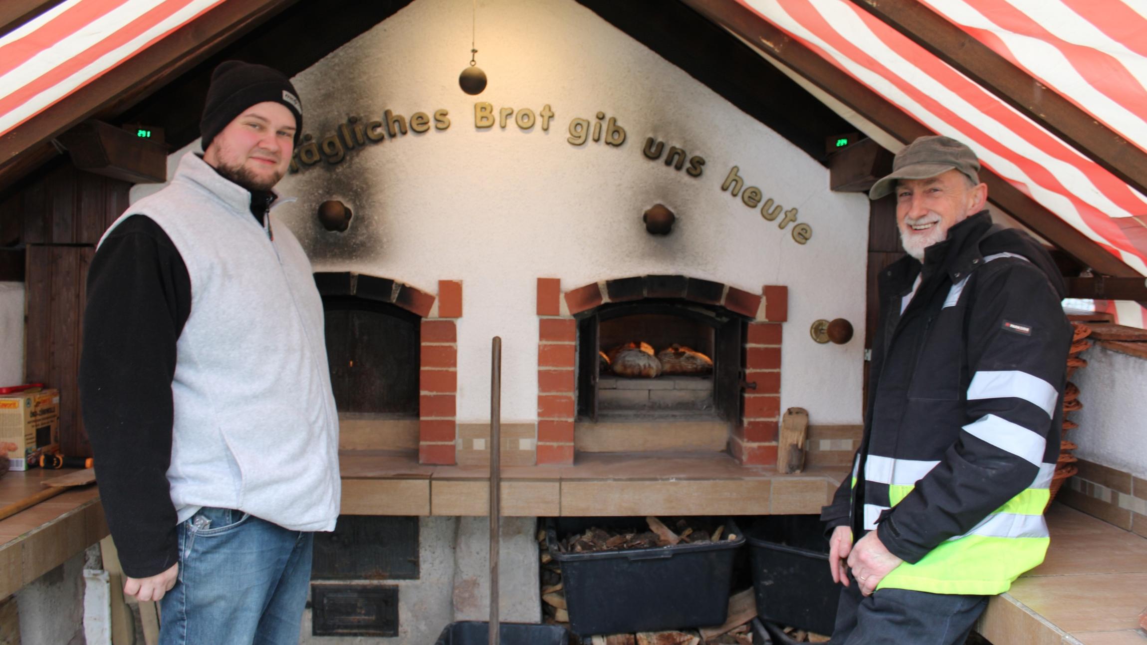 In einem überdachten Außenbereich stehen zwei Personen vor einem großen, gemauerten Holzbackofen. Der Ofen besitzt zwei Backkammern, in denen Brotlaibe auf Steinflächen liegen und backen. Über dem Ofen ist der Schriftzug „Tägliches Brot gib uns heute“ angebracht. Unterhalb der Arbeitsfläche stehen mehrere schwarze Kunststoffwannen mit Holzstücken. Das Dach besteht aus schräg verlaufenden Balken und ist mit einer rot-weiß gestreiften Plane überspannt.