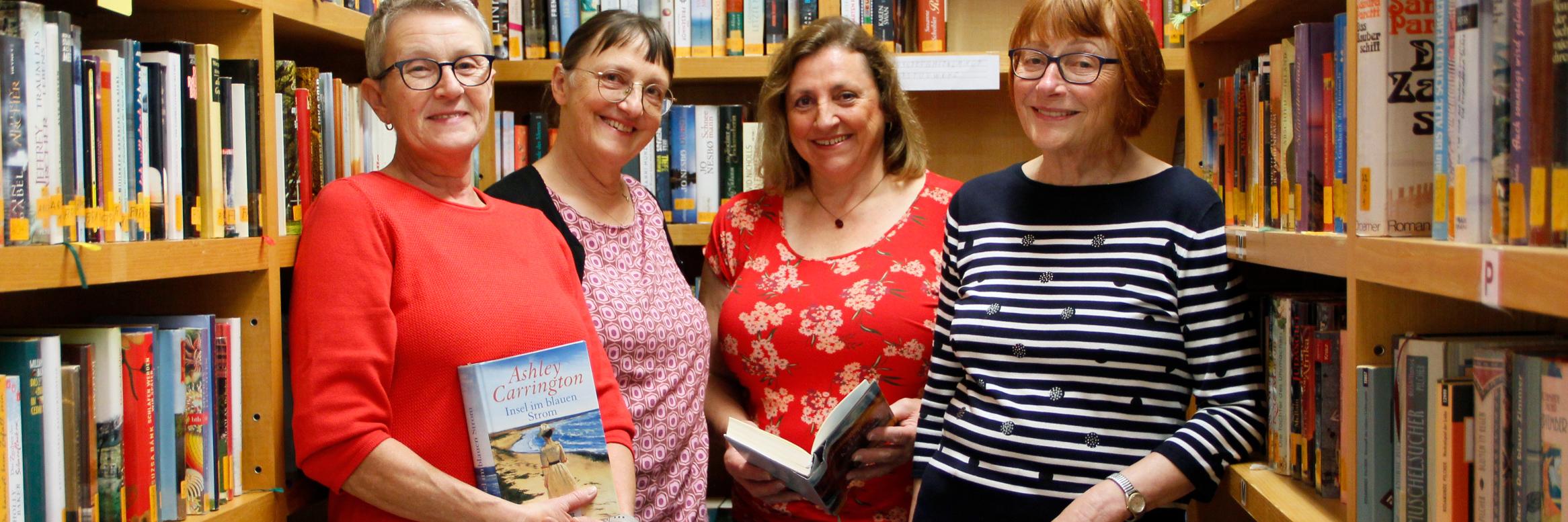 Vier lächelnde, etwas ältere Damen stehen vor Bücherwänden, zwei haben Bücher in der Hand.